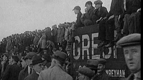 British Pathe Archive still of hundreds of people awaiting news at the scene of the tragedy in 1934. People sit on coal trucks and stand together looking off at the site of the explosion
