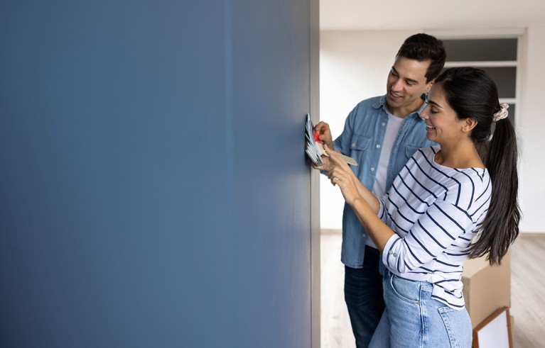 A happy couple hold up a set of paint colour cards against a blue wall in their home.