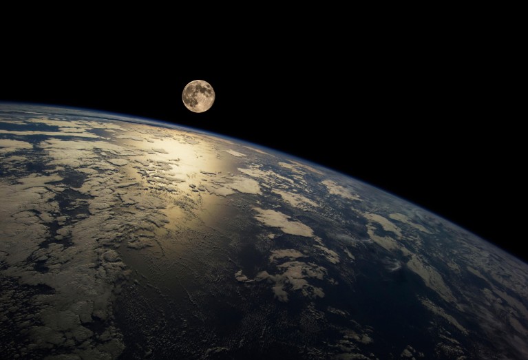 A view from Earth's orbit of the moon hovering over the curved edge of Earth surrounded by a thin layer of atmosphere visible as a blue haze