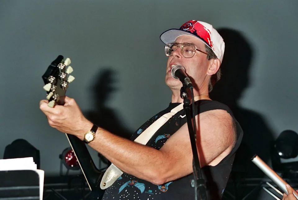 a man with a baseball cap and glasses plays guitar and sings into a microphone