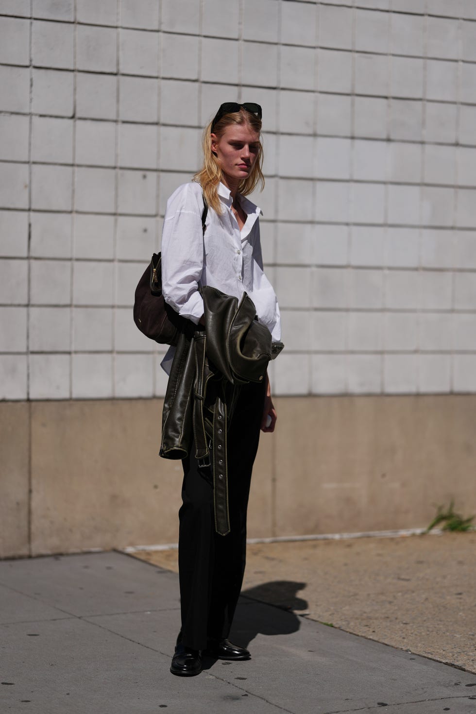new york, new york september 11: a guest wears shoulder length blonde hair worn down, black rectangular sunglasses pushed up on the head, a dark brown suede crescent hobo bag with a zipper and gold tone hardware on the shoulder, an oversized white cotton button up shirt with a spread collar and button cuffs, a dark olive leather biker jacket with a belted hem and metal grommets carried over the arm, black straight leg trousers, and black polished leather loafers with low heels, outside michael kors, during new york fashion week, on september 11, 2025 in new york, new york (photo by edward berthelot/getty images)