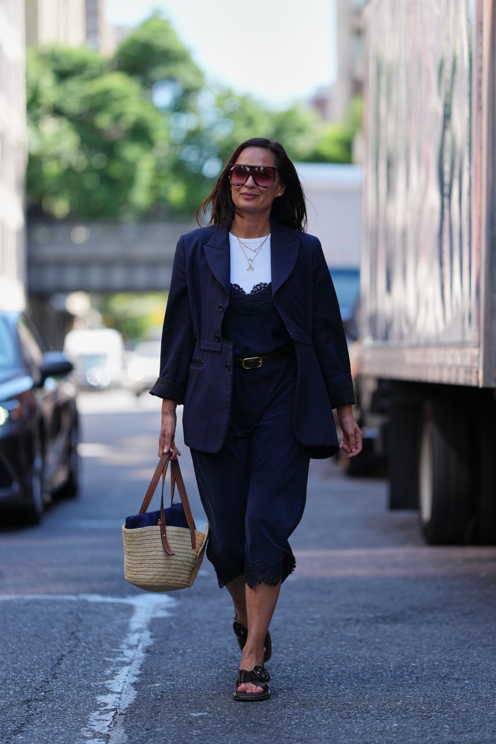 new york, new york september 11: roopal patel wears dark brown straight hair worn loose, oversized rectangular sunglasses with brown tinted lenses, layered gold necklaces, a woven straw basket bag with tan leather top handles, a navy blazer jacket with notch lapels and rolled sleeves over a white crew neck t shirt and a navy satin slip dress with lace trim at the neckline and hem, cinched with a black leather belt with a gold tone buckle, black leather slide sandals with cutout uppers and flat soles, outside lafayette 148, during new york fashion week, on september 11, 2025 in new york, new york (photo by edward berthelot/getty images)