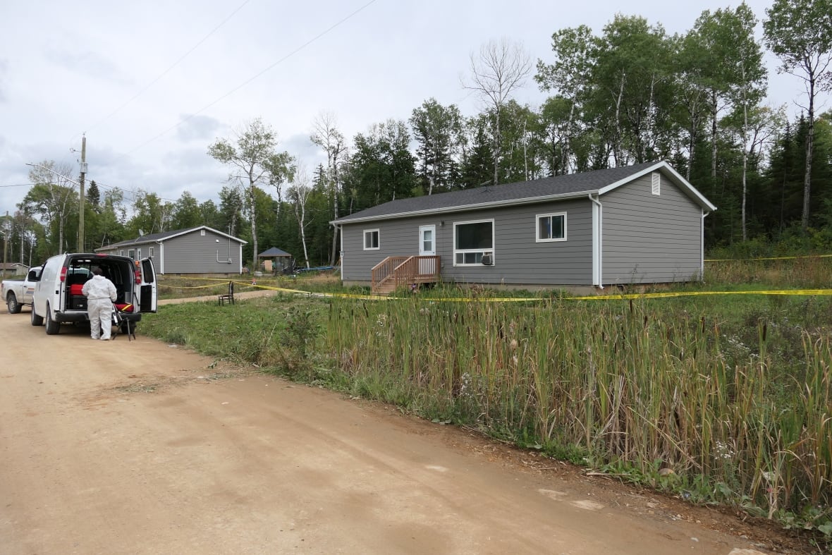 An investigator in white coveralls stands behind a white van parked in front of a grey house surrounded by police tape.