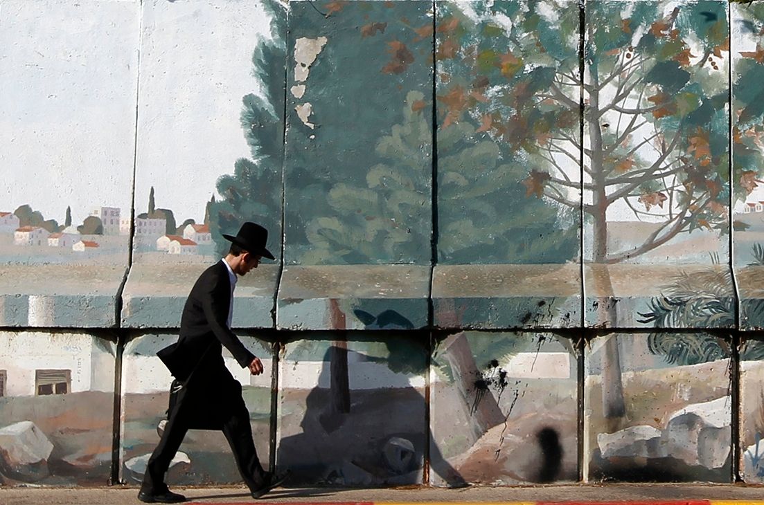 An ultra-Orthodox Jewish man walks along a concrete barrier painted with a mural depicting a serene landscape with trees and buildings.