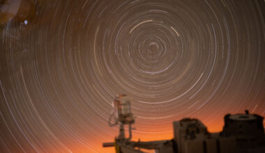 Stars photographed over 31 minutes make a concentric pattern of white circles. At the bottom edge of the photo, there is a soft orange glow. A portion of the International Space Station can be seen in the foreground. This photo was taken through a window on the space station.