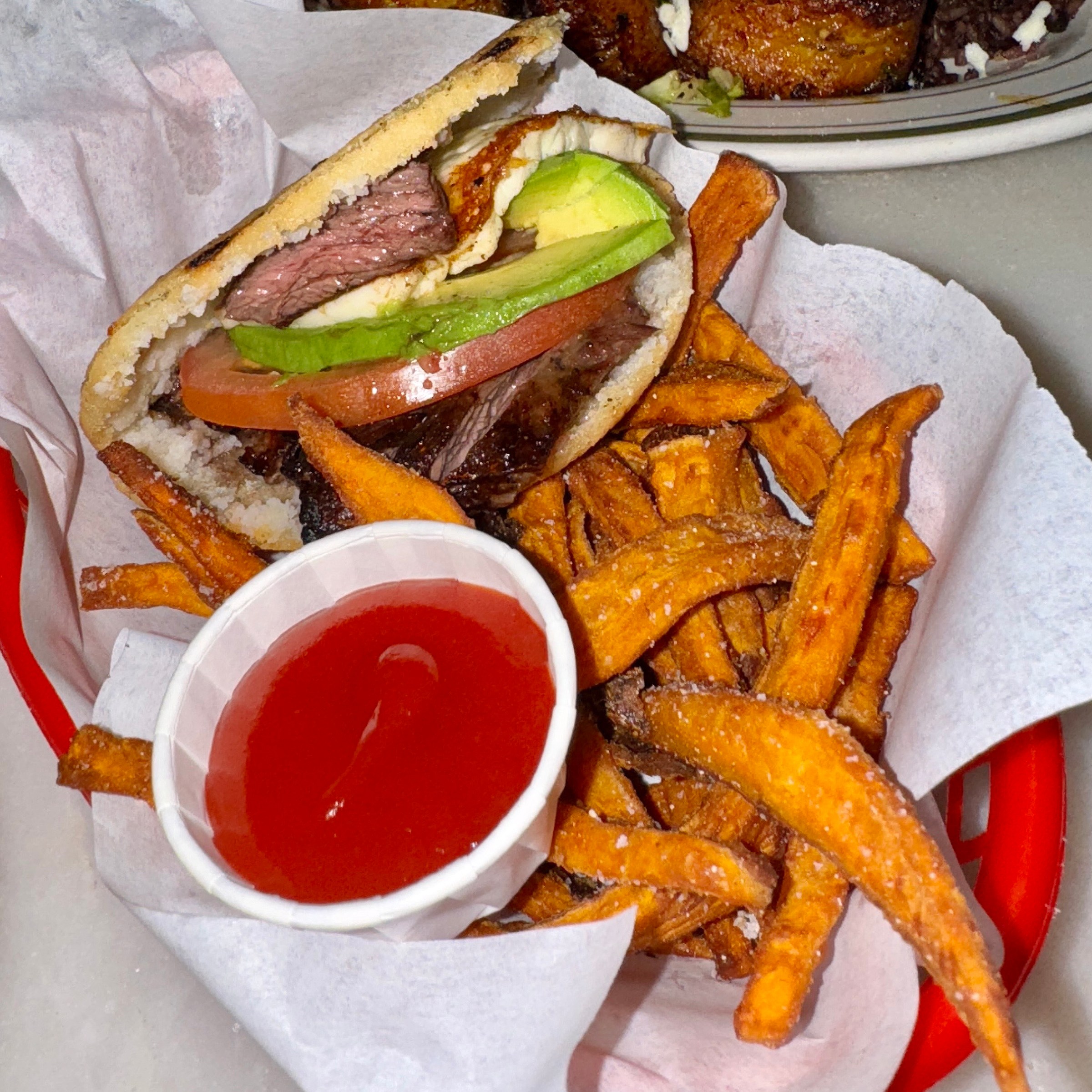A sandwich and fried on a wax paper on top of a red tray with some red sauce in a paper container.