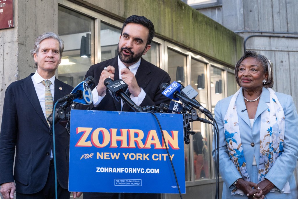 Zohran Mamdani speaks at a podium with microphones, flanked by New York State Senate Majority Leader Andrea Stewart-Cousins and another man.