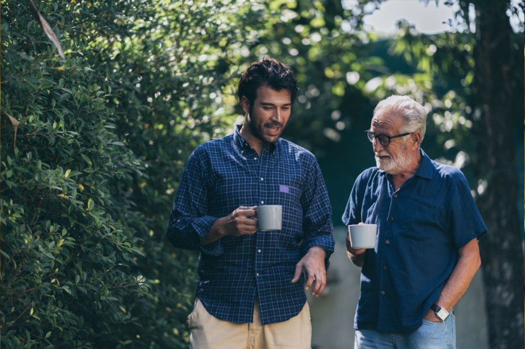 A younger man walking with an older man drinking coffee and talking