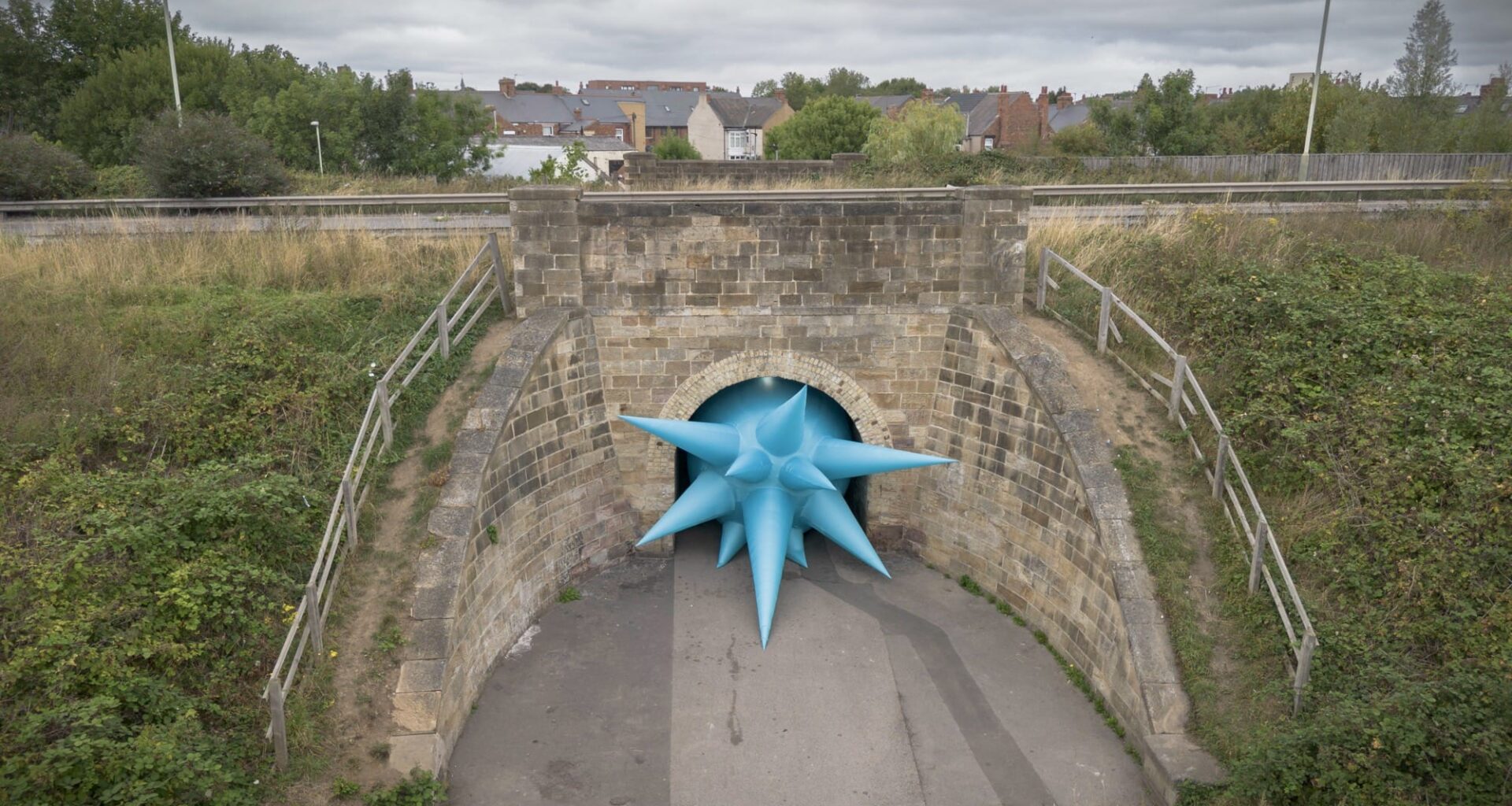 an aerial view of a large-scale installation by Steve Messam under an old stone bridge of a blue, spiked form that appears wedged in the archway under the bridge