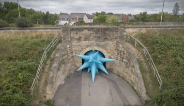 an aerial view of a large-scale installation by Steve Messam under an old stone bridge of a blue, spiked form that appears wedged in the archway under the bridge