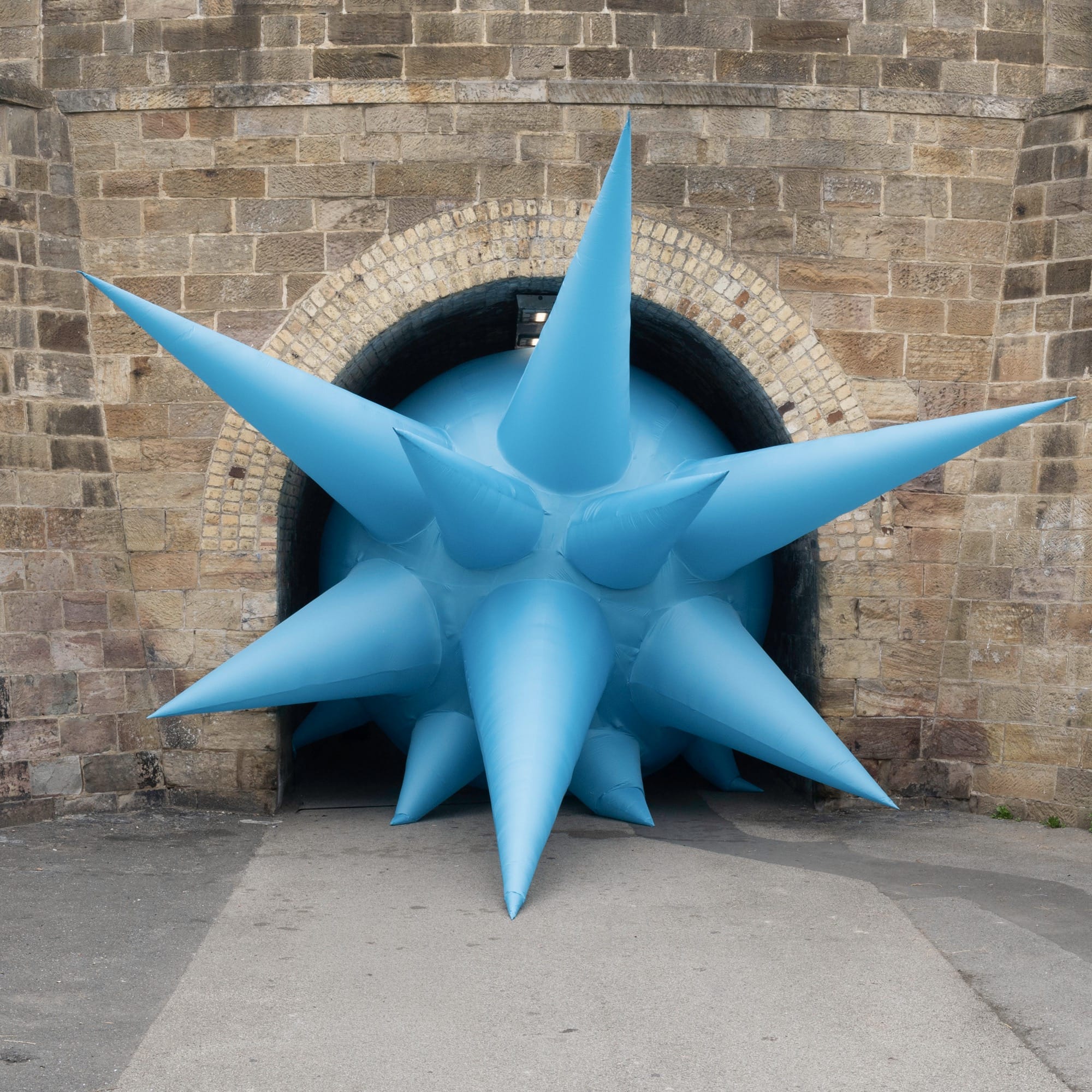 a large-scale installation by Steve Messam under an old stone bridge of a blue, spiked form that appears wedged in the archway under the bridge