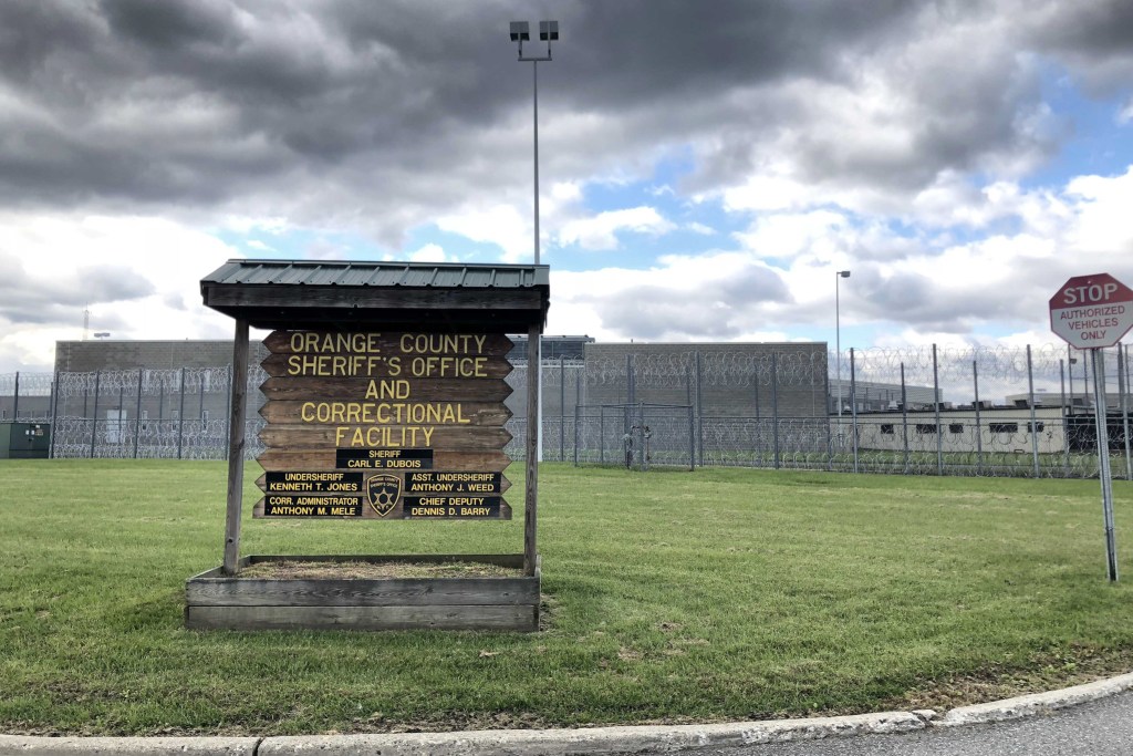 "Orange County Sheriff's Office and Correctional Facility" read a wooden sign in front of a barbed wire fence surrounding a prison building.