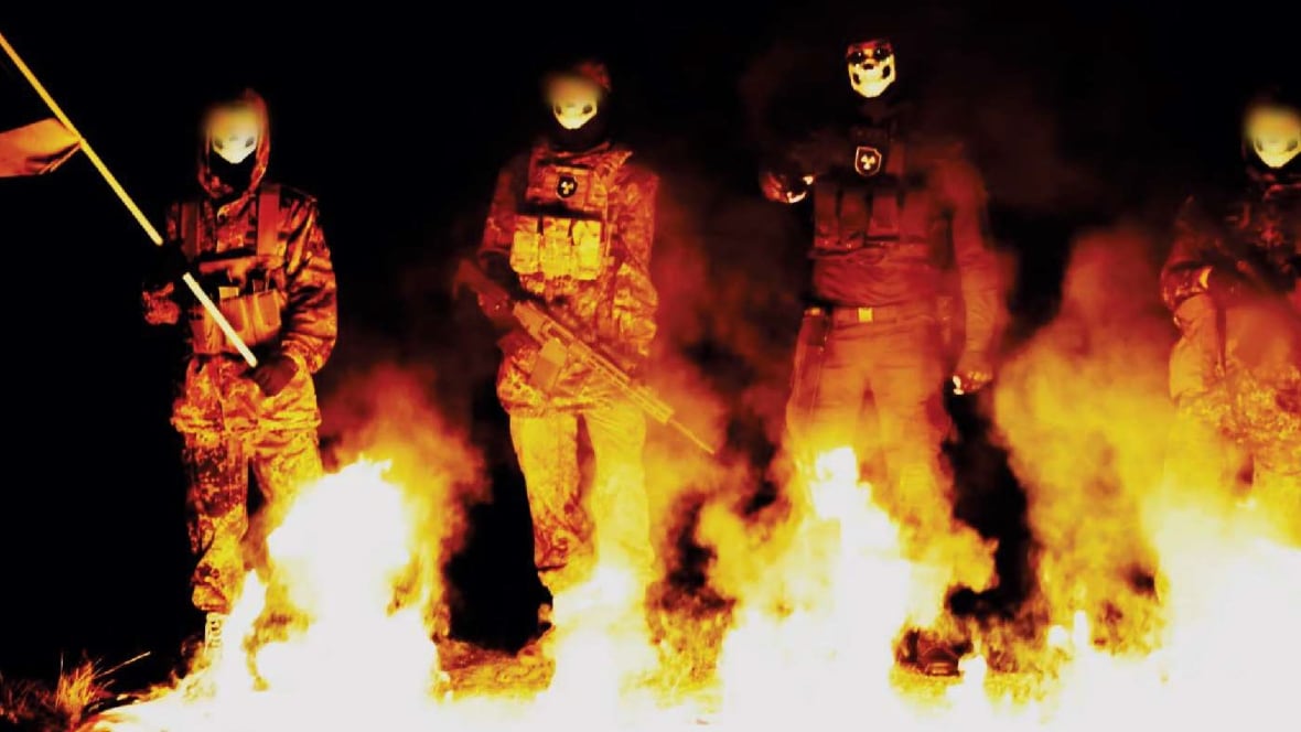 A group of men in masks and combat fatigues stand in front of a fire.
