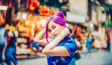 A young woman with vibrant purple hair, wearing a blue patterned dress, poses with her hands near her head on a bustling, colorful city street with blurred people and lights in the background.