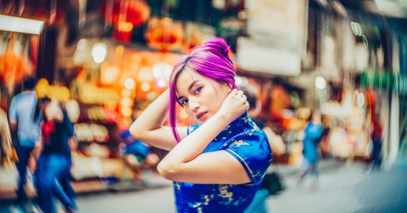 A young woman with vibrant purple hair, wearing a blue patterned dress, poses with her hands near her head on a bustling, colorful city street with blurred people and lights in the background.