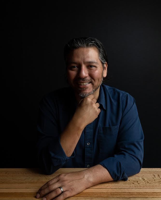 A chef smiles while holding a hand under his chin at a wooden table.