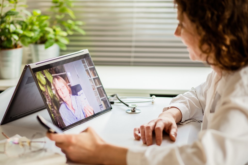 A woman having a telemedicine consultation with an elderly woman visible on her laptop screen.