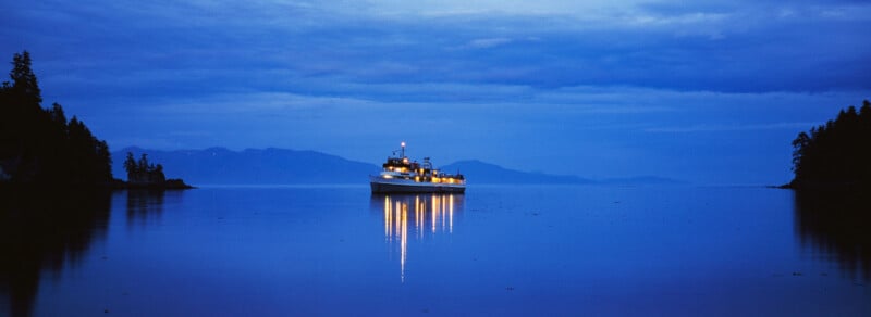 A boat with lights reflecting on calm blue water is anchored between tree-lined shores at dusk, with distant mountains under a cloudy sky.