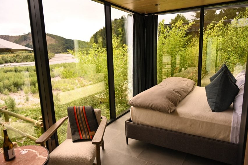 Glass cabin interior with a double bed, side table and chair overlooking a river