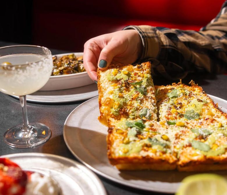 A hand pulling a slice from a Detroit-style pizza with a cocktail on the table.