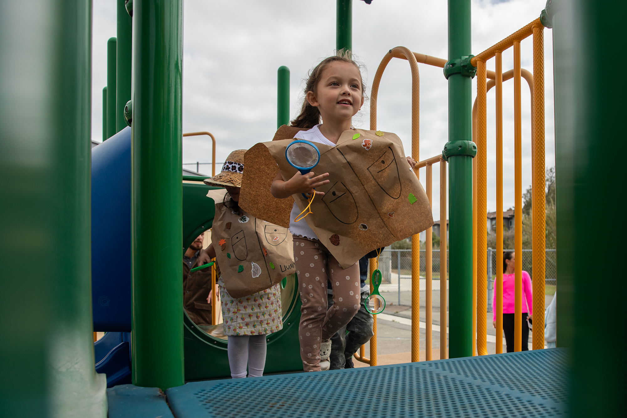 A young student — wearing a brown safari vest made out of construction paper — runs up a jungle gym as they hold a magnifying glass during a school activity.
