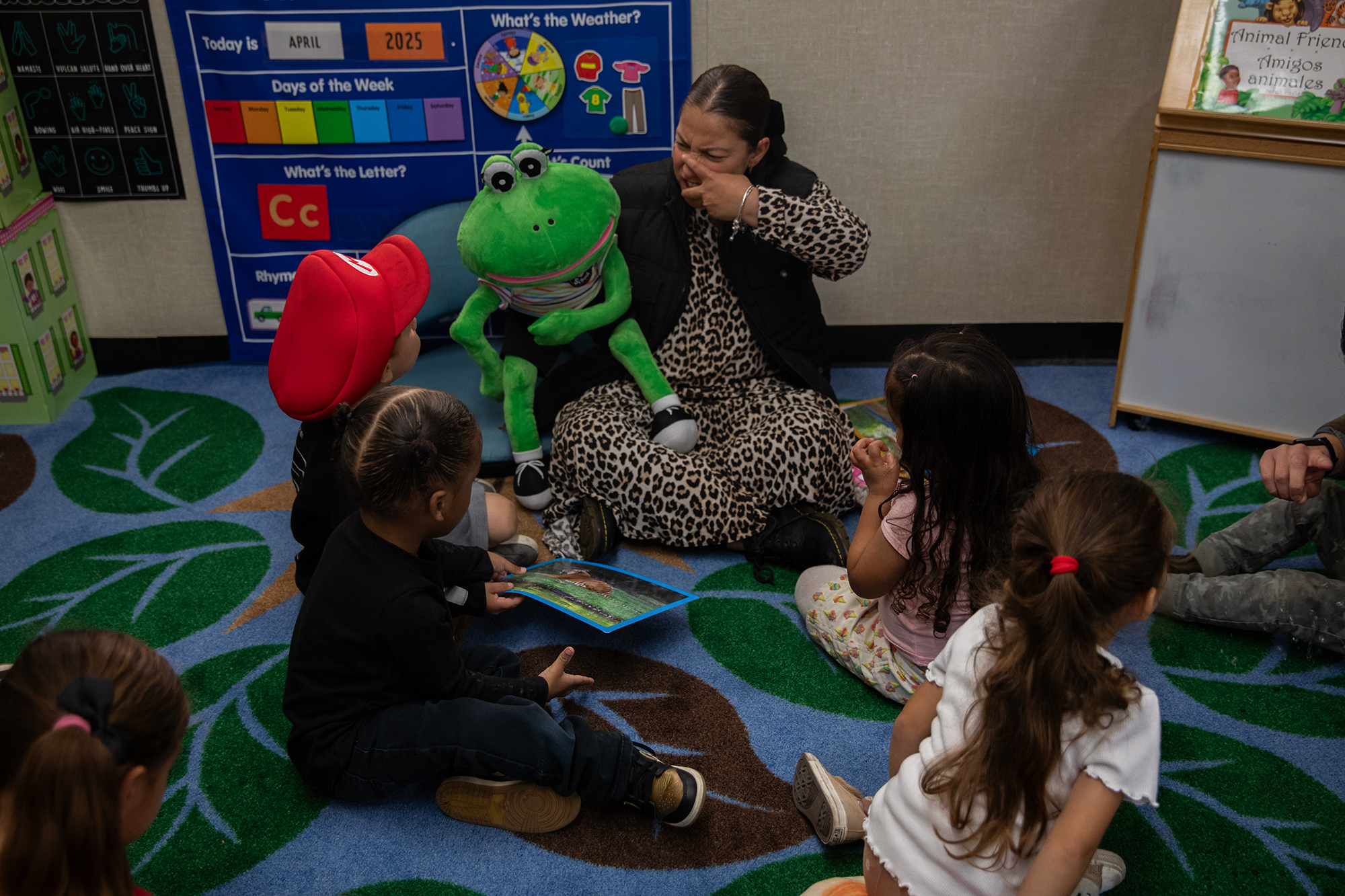 A teacher — wearing a leopard-print dress and a black puffy vest — holds a frog puppet to teach students about different animals as they all sit on a colorful rug inside a classroom.