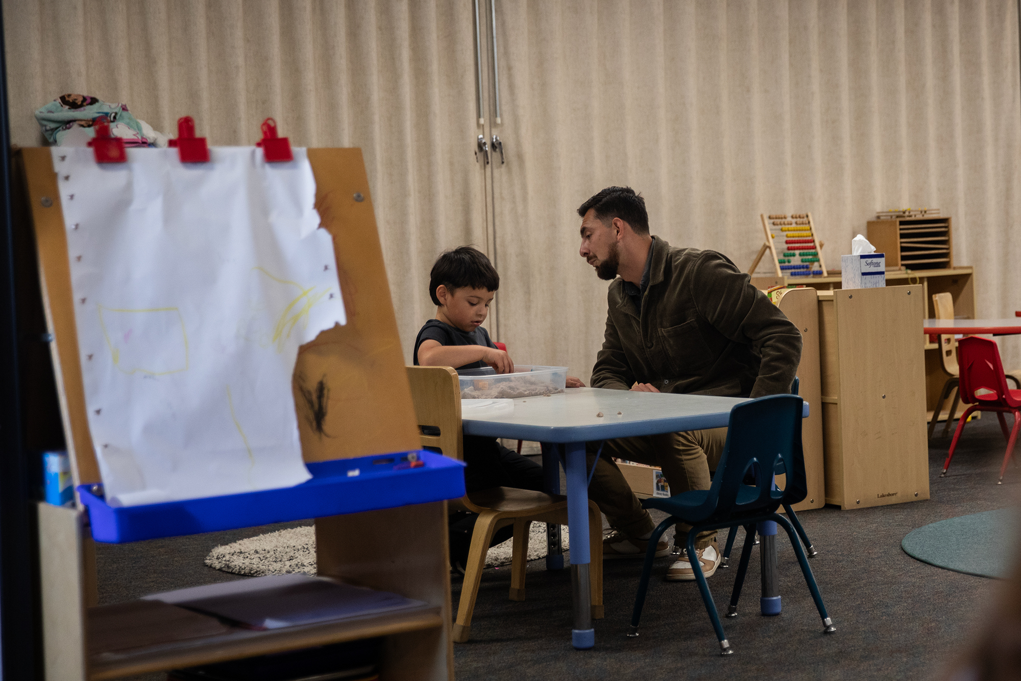 An adult sits next to a child as hey play with a box filled with sand, inside a classroom.