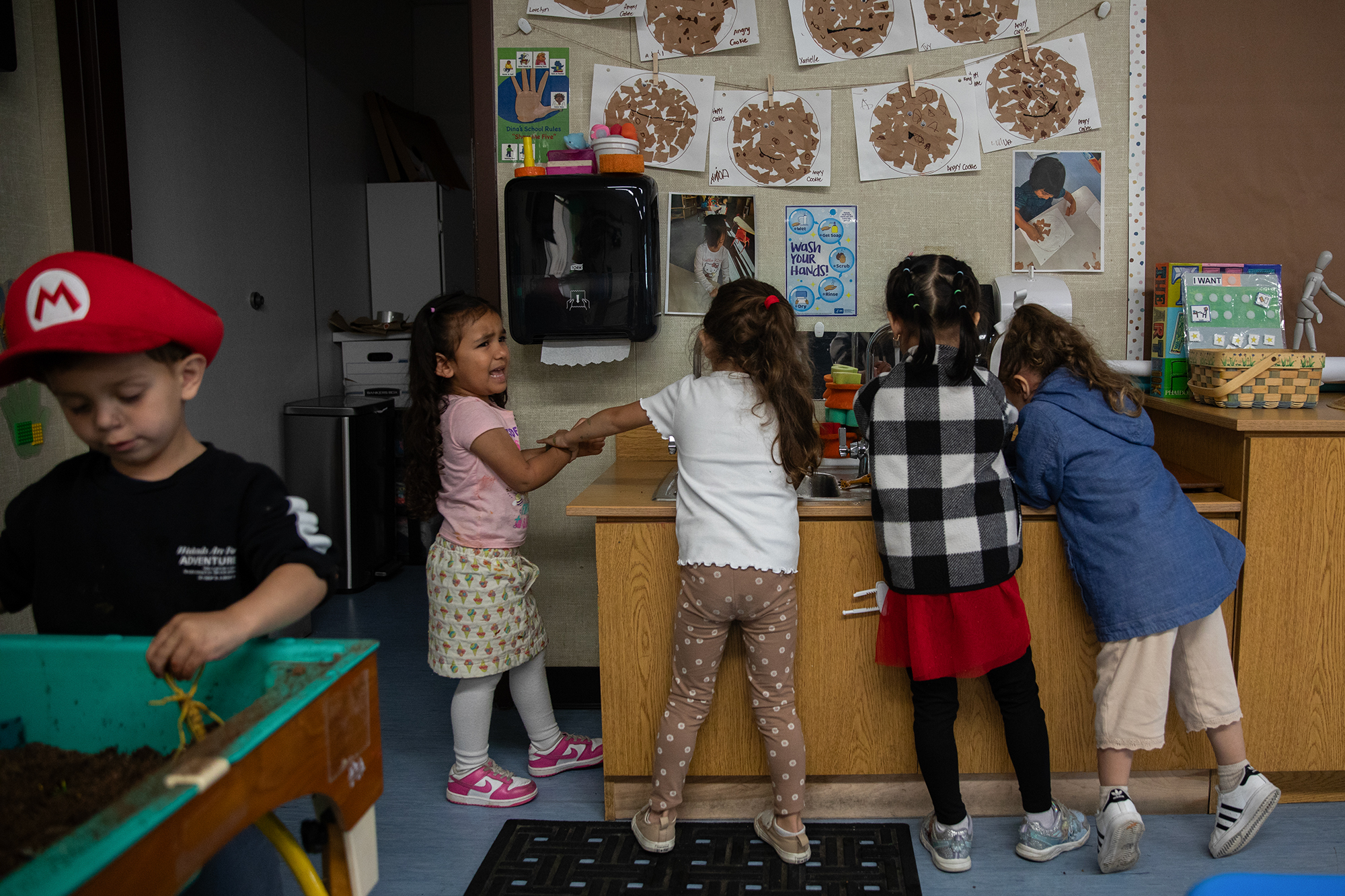 Young students stand in front of a sink inside a classroom as they wash their hands after a painting activity. One student, on the far left, cries as another grabs their hand.