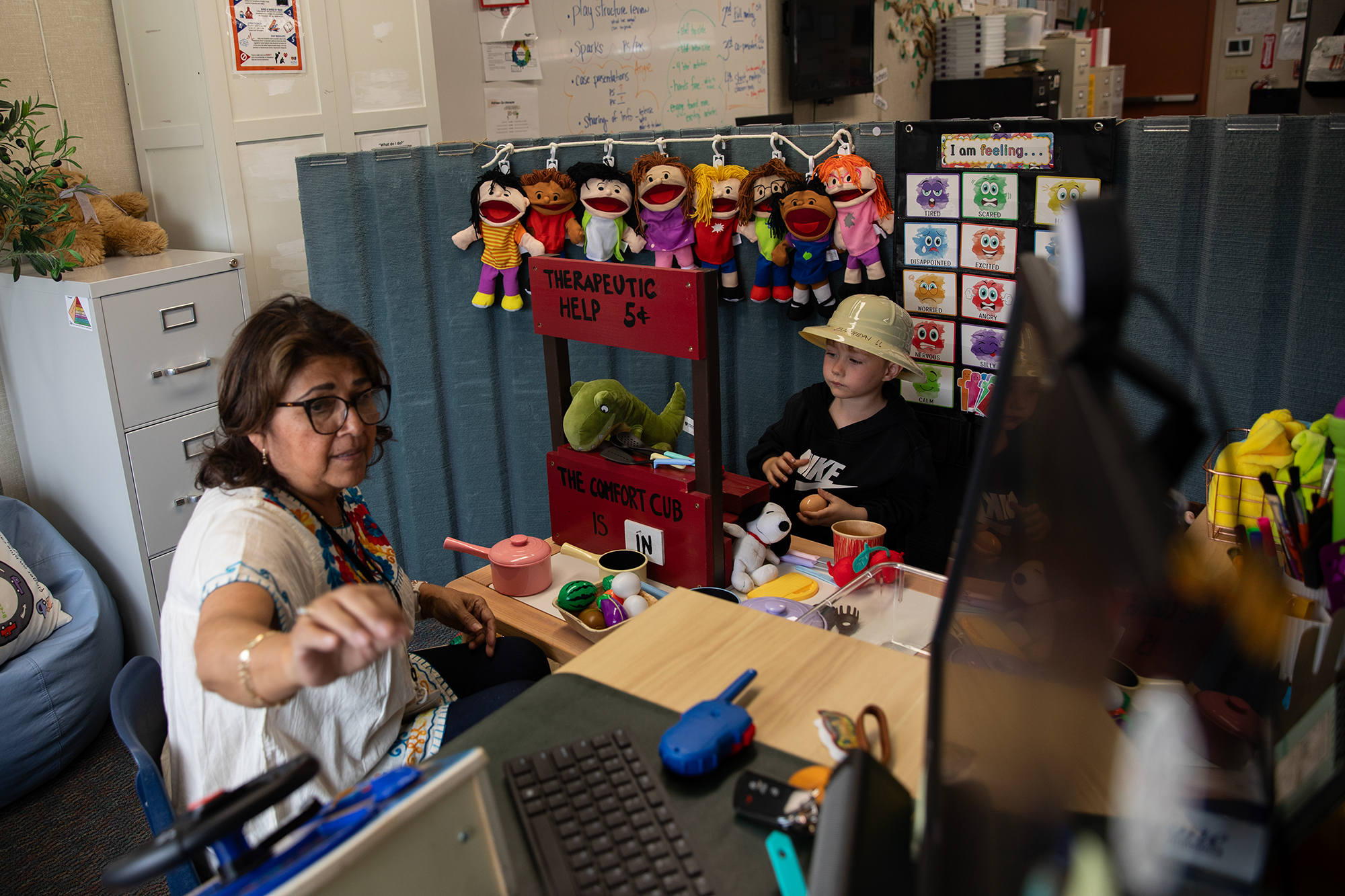 An adult reaches to grab something on their desk while they play with a child during a therapy session. The child, wearing a plastic safari hat, stands in front of a small red wooden stand that reads 'therapeutic help" as they are surrounded by toys and puppets.