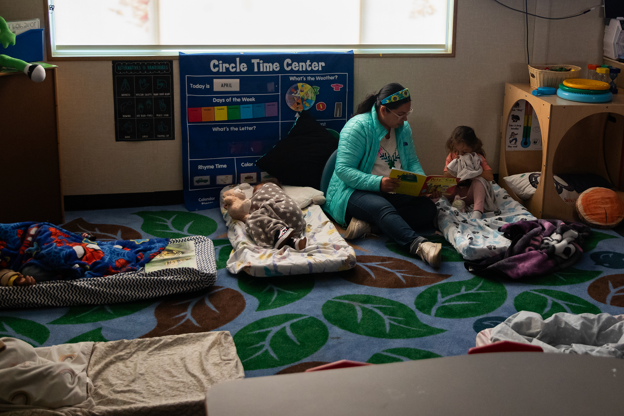 A teacher reads to a young student who struggles to sleep during nap time. Other students can be seen sleeping nearby as they all on small napping mats.