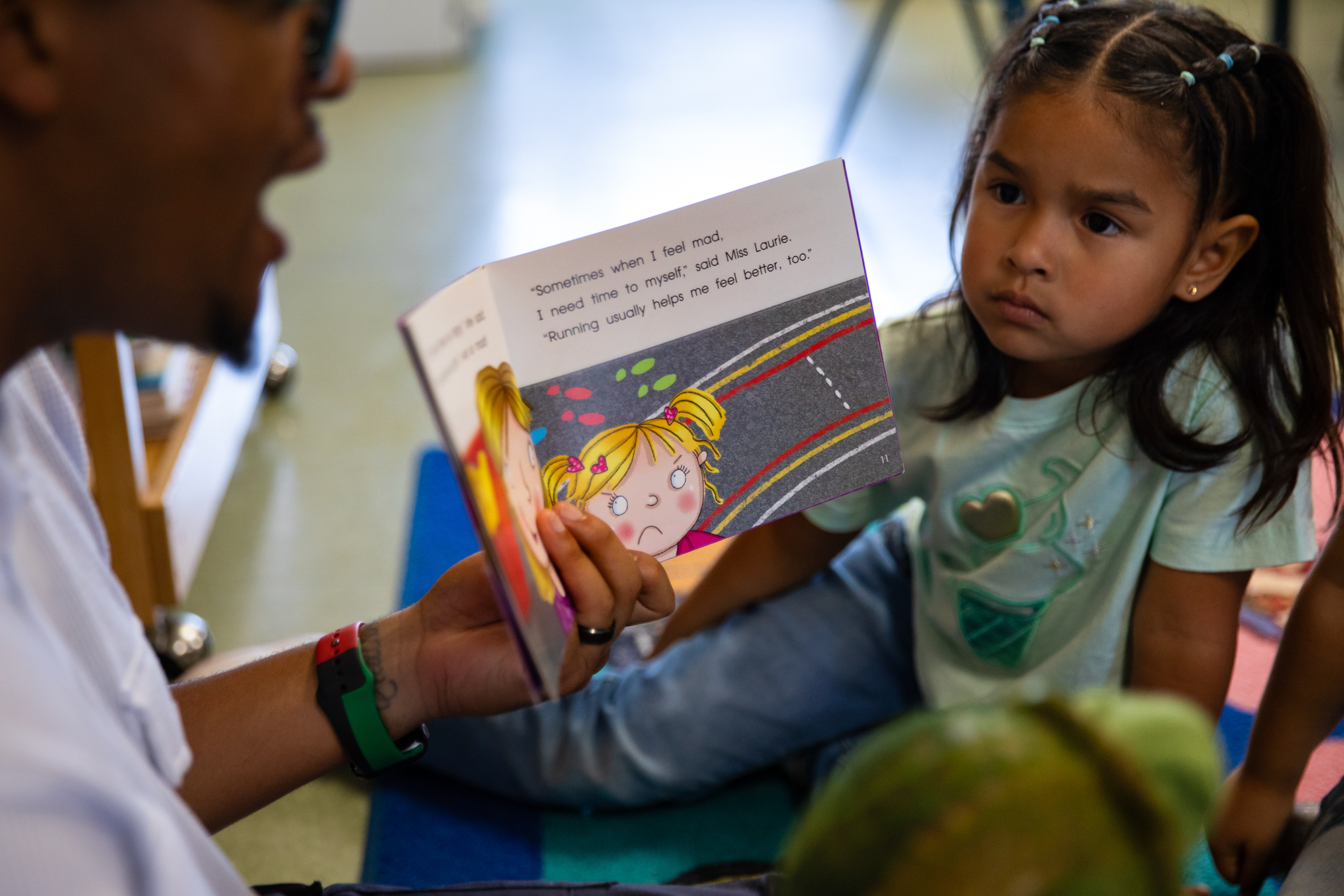 A young student listens as a teacher holds and reads a book to a small group of students. A page of the book shows an illustration of a young girl and reads "'Sometimes when I feel mad. I need time to myself'", said Miss Laurie.Running usually helps me feel better, too."