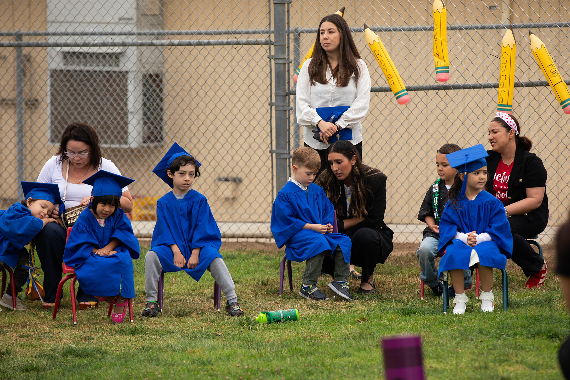 A school officials leans down and talks to a student who looks towards the ground as they sit next to other graduates. The students wear blue graduation gowns and caps.