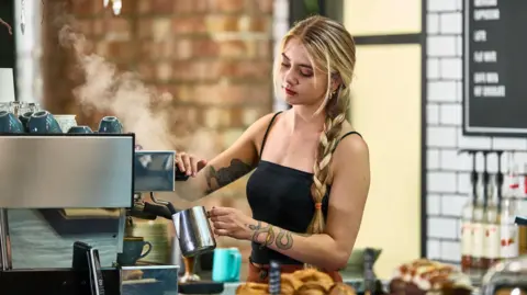 Getty Images A young woman barista with long blonde hair in a plait wearing a black vest top making a coffee behind a counter with pastries in the foreground and syrups in the background