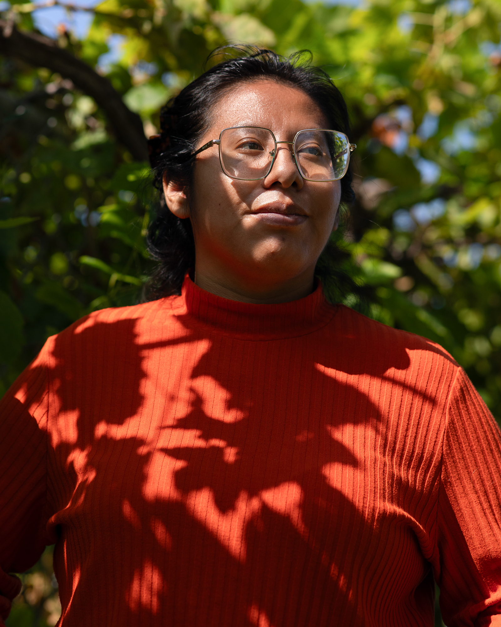A person — with long black hair tied-up in a ponytail and wearing reading glasses and an orange turtle neck — looks to their right as they stand in front of a wall with green ivy vines on it.