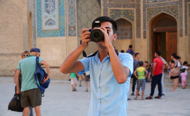 A young man in a light blue shirt takes a photo with a camera in a busy outdoor area with historic, ornate buildings in the background and people walking around.