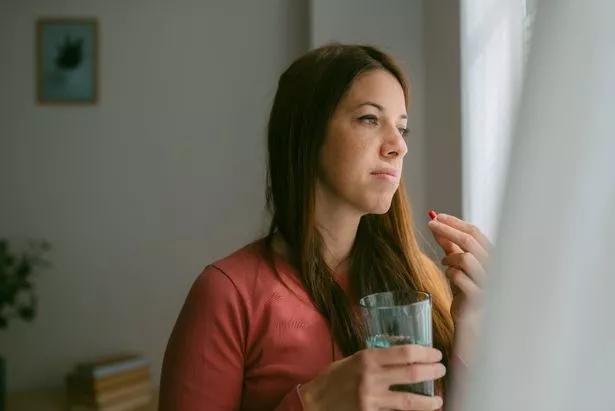 Portrait of a young woman taking a capsule of vitamin pills standing by the living room window. Caucasian woman, brunette. She is wearing casual clothes. Image with copy space on the left.