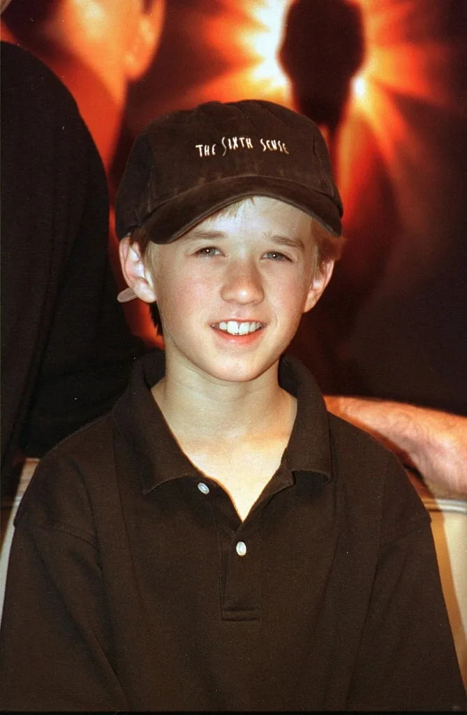 A young actor wearing a cap with "The Sixth Sense" logo, smiling at an event