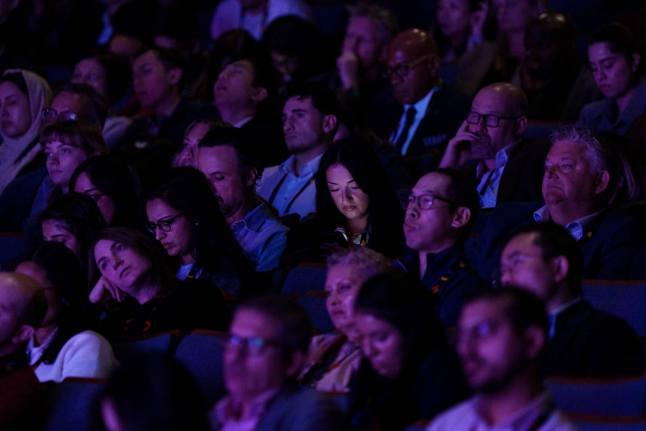 Seated in an audience of people, a woman's face is lit up by the glow of her phone screen.