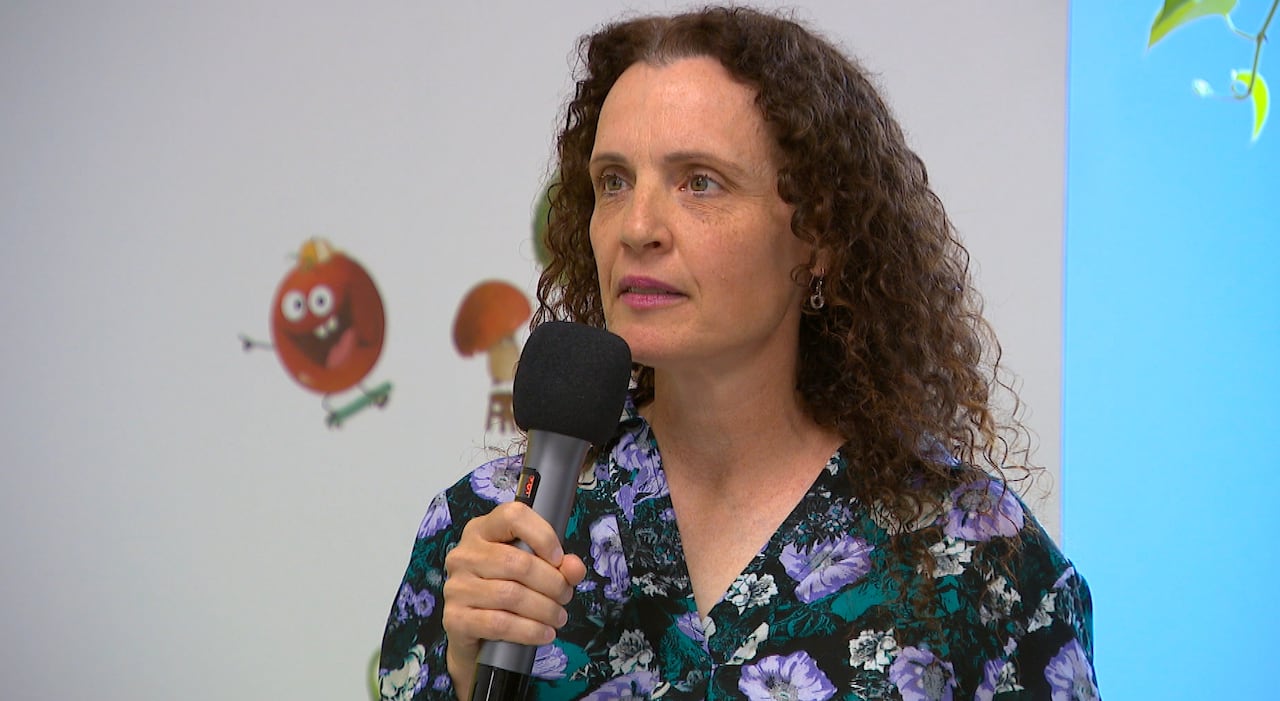 A woman with long, curly brown hair speaks with a microphone. 
