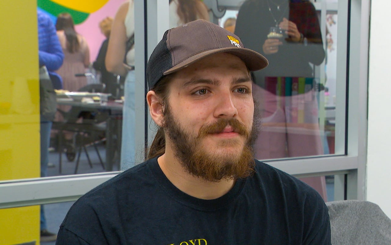A young man with a beard and wearing a ball cap sits at a table and speaks into a microphone.