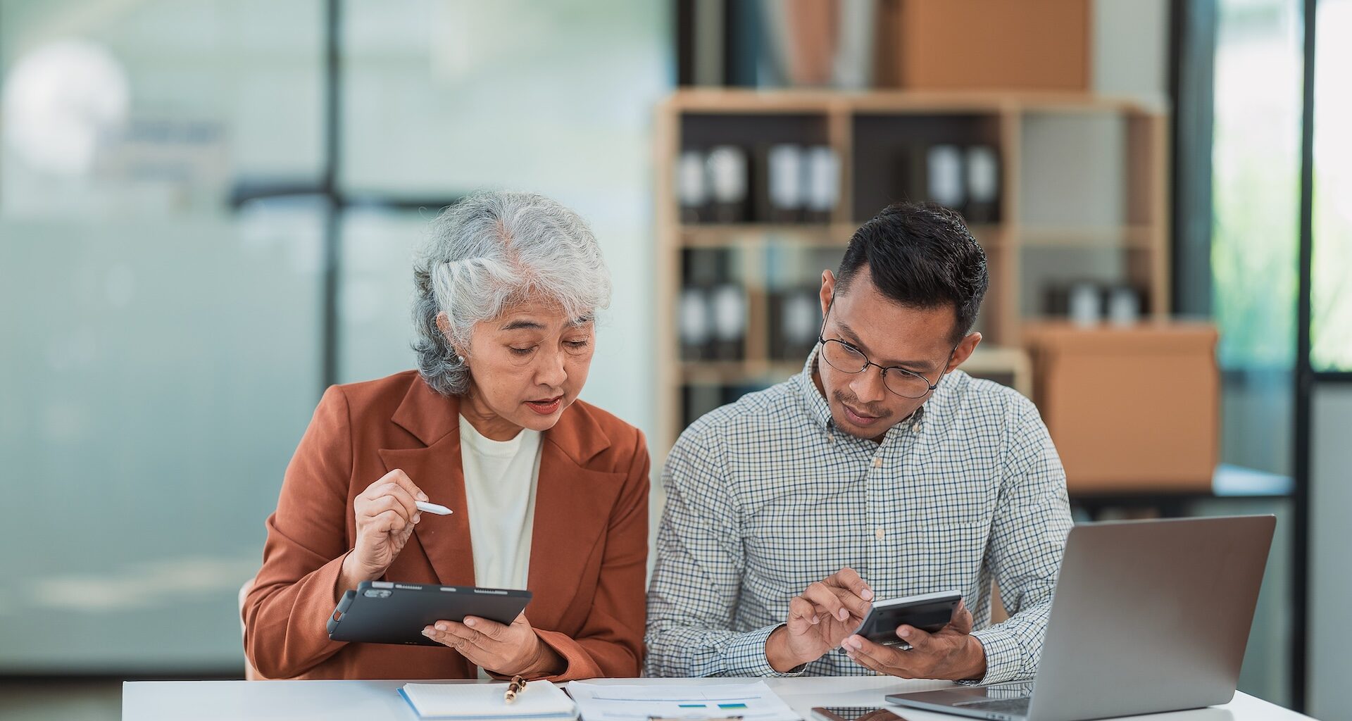 Senior Asian business woman and young man working together analyzing finances senior staff teach marketing and planning to younger staff woman holding graph and chart with a happy smile.