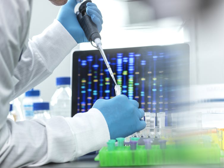 Person in white coat pipetting a sample into a vial, a DNA readout on a screen in the backdrop