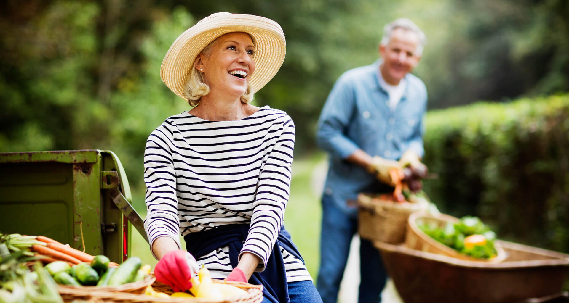 Portrait of mature woman with baskets of harvested vegetables and man working in the background.