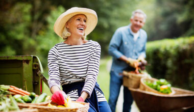 Portrait of mature woman with baskets of harvested vegetables and man working in the background.