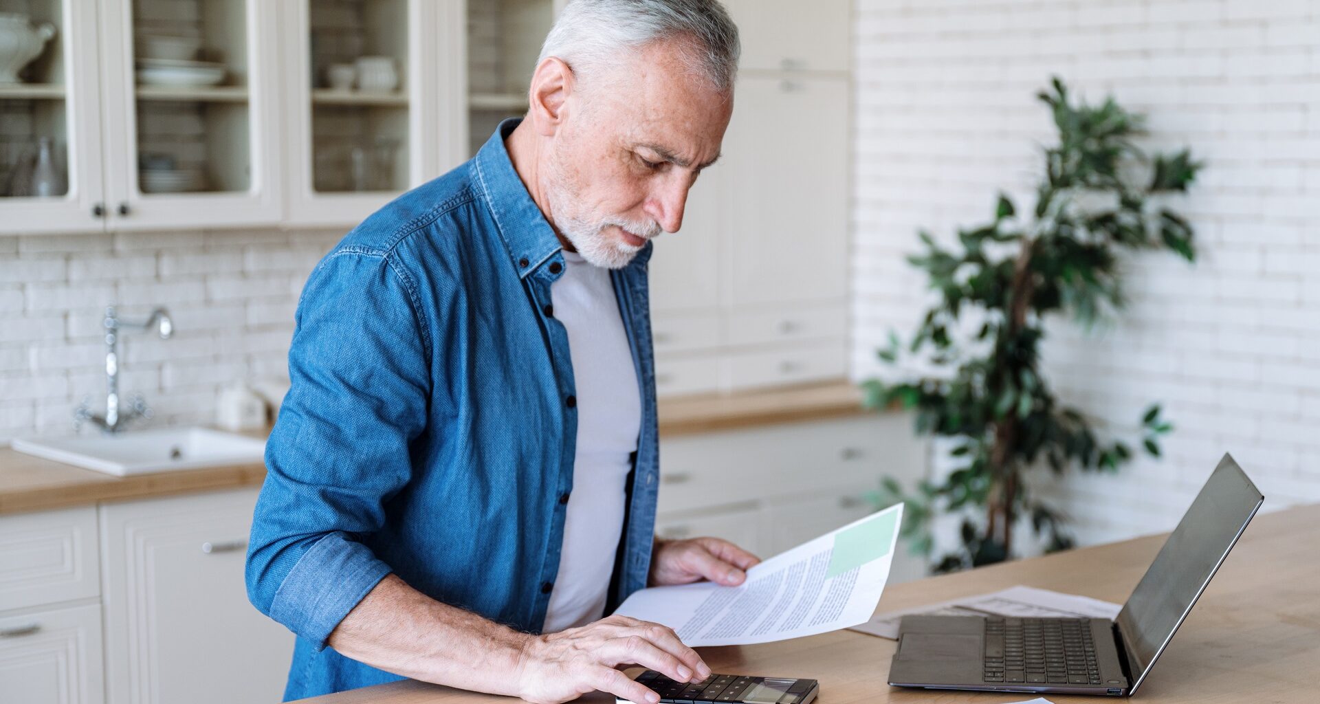Focused senior man using a calculator and a laptop.