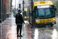 A man waits for a DART bus along Commerce Street as heavy rain falls in downtown Dallas,...