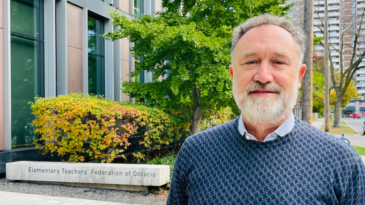 A bearded man in a blue sweater stands outside a building, with a low stone sign denoting Elementary Teachers' Federation of Ontario seen behind.