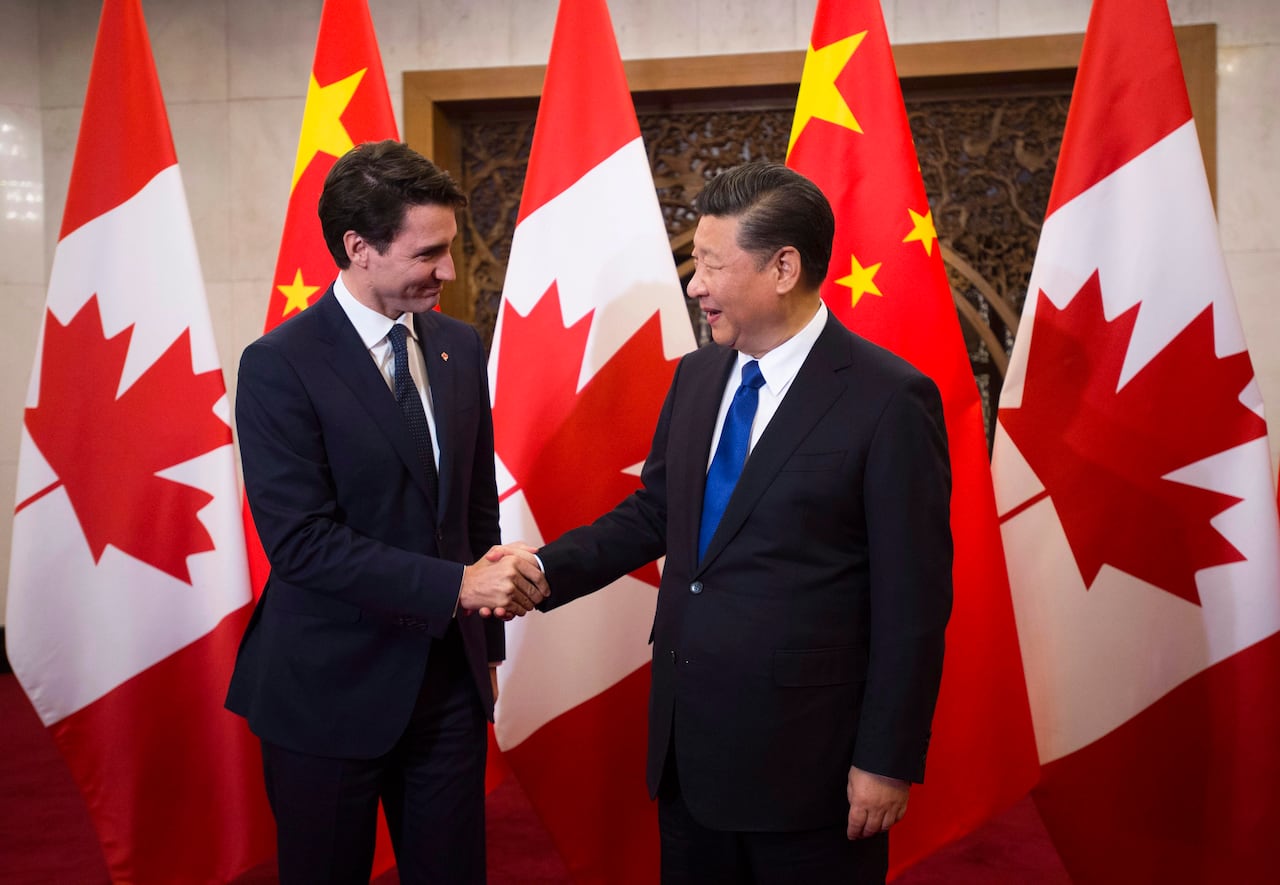 Two men in suits are shaking hands in front of a row of Canadian flags.