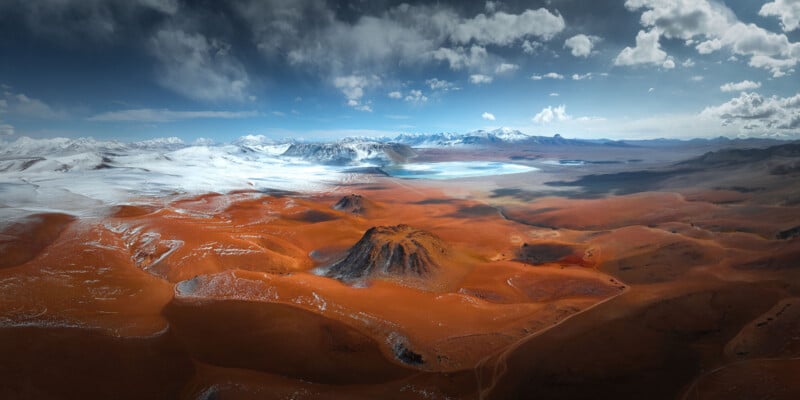A dramatic landscape showing red-orange desert hills, a dark central hill, distant snowy mountains, and a blue-white lake under a partly cloudy sky.