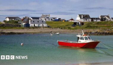 A small fishing boat with a red hull is tied up in a shallow expanse of turquoise water. Land in the background is dotted with white houses.